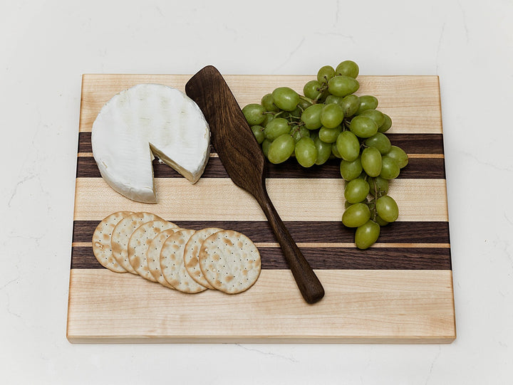 maple and walnut edge grain cutting board with cheese, crackers, and grapes on a white background