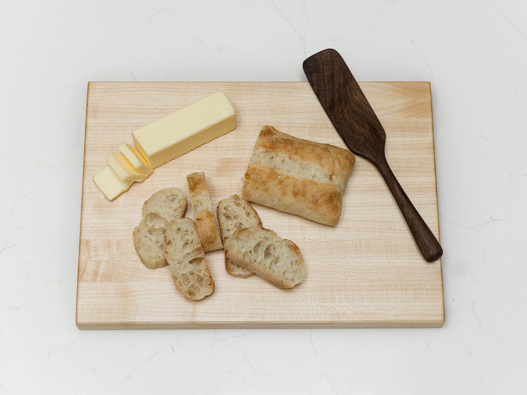 maple edge grain cutting board with sliced bread, a stick of butter, and a wooden spatula on a light background