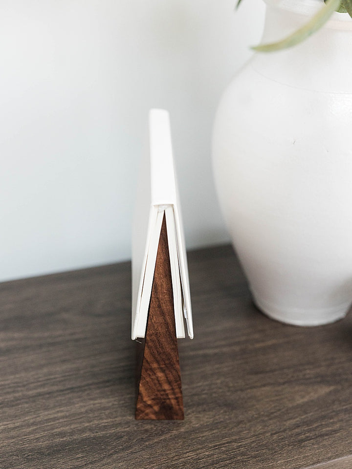 Solid walnut book wedge with white journal on top sitting wood table with a white vase in the background