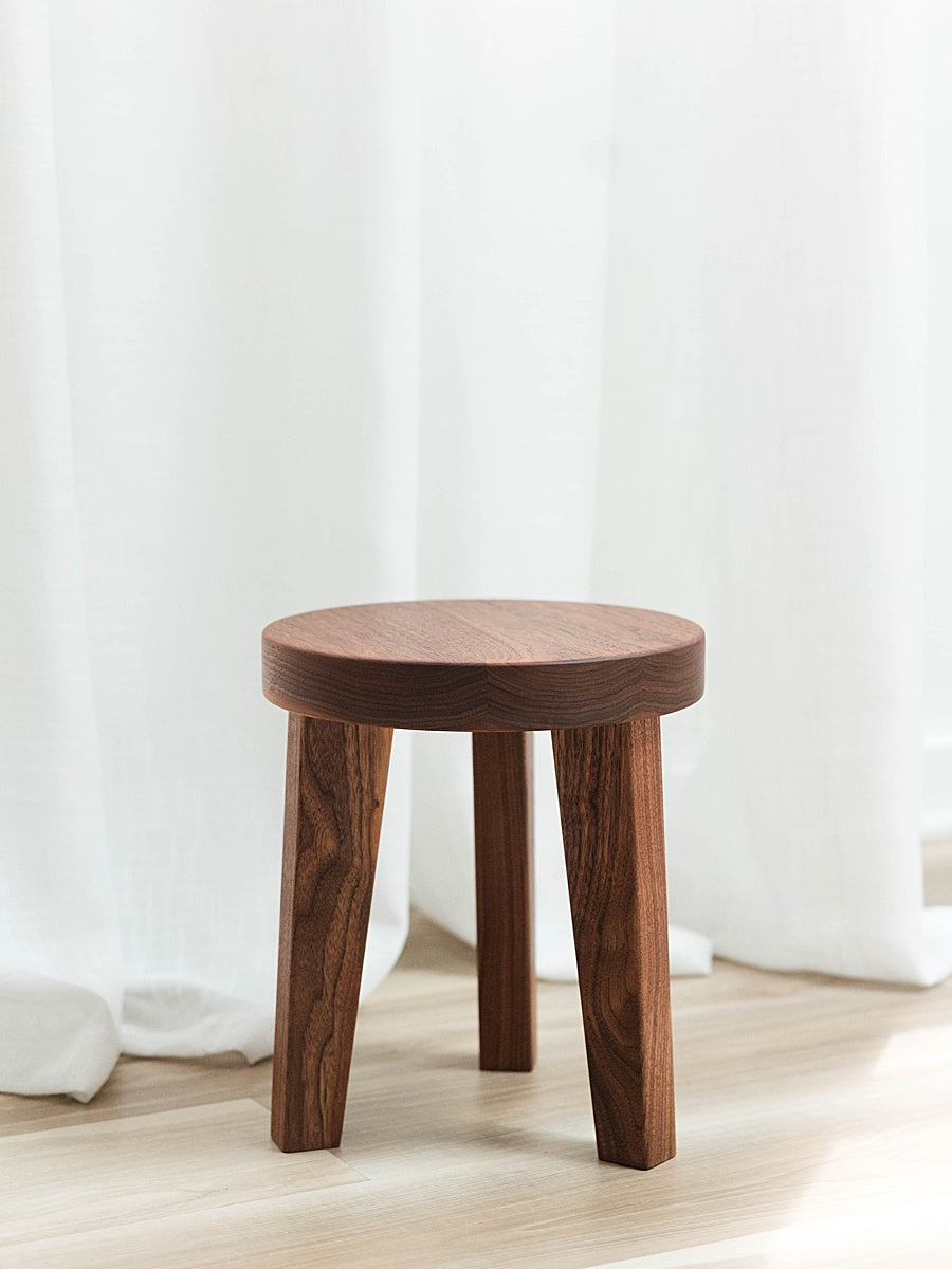 Solid walnut stool with a round top and three legs against a white curtain and light colored floor