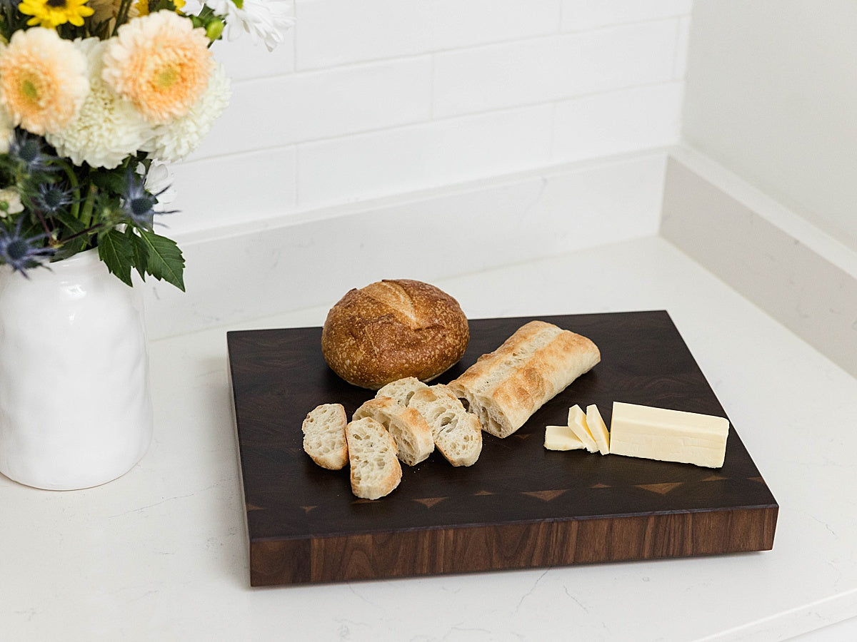 Walnut end grain cutting board with bread and butter on a white surface with flowers in the background