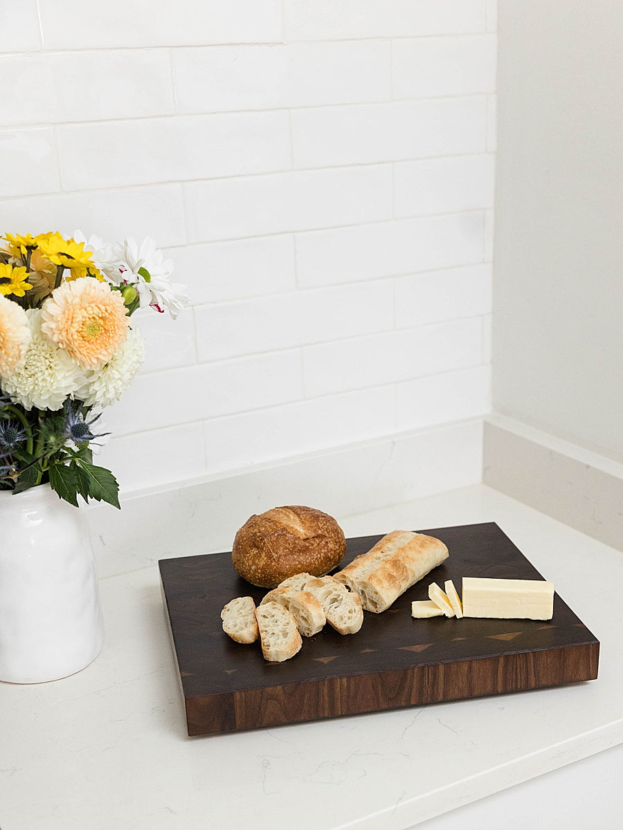 Walnut end grain cutting board with bread and butter on a white surface with flowers in the background