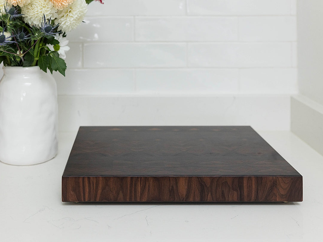 Dark walnut end grain cutting board on a white surface with a white vase of flowers in the background.