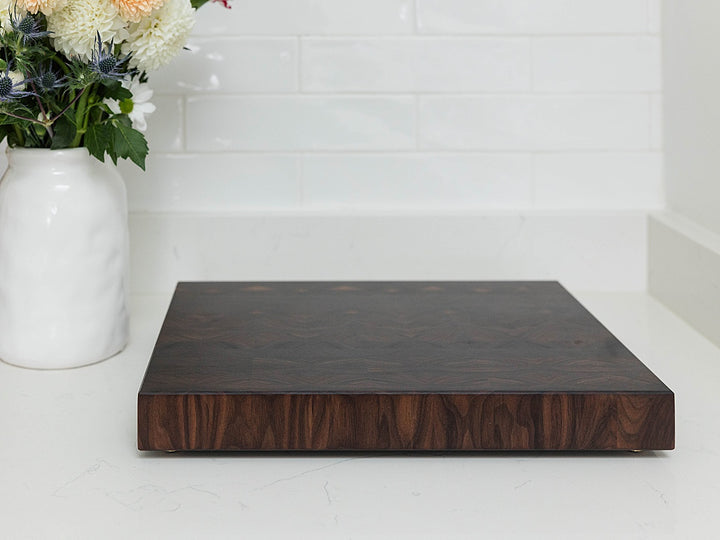 Dark walnut end grain cutting board on a white surface with a white vase of flowers in the background.