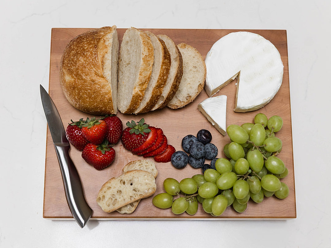 Wooden maple end grain cutting board with bread, cheese, strawberries, grapes, and blueberries on a white background