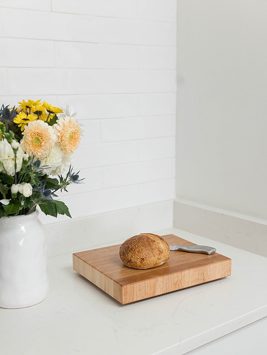 Wooden cutting board with a loaf of bread on a white surface, next to a vase of flowers.